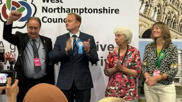 Two men and two women on stage. They are all clapping except one man, who is pumping the air.