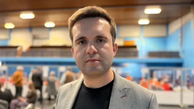 A man with short brown hair, a black tshirt and a grey jacket in a room with lots of people stood behind him