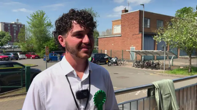 A man with dark curly hair, a pale shirt and a green rosette standing in front of metal railings with a large brick building in the background
