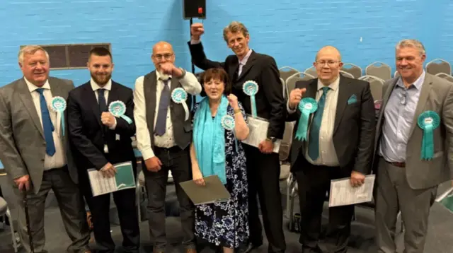A gathering of men in suits and one woman in a dress, some with their fists in the air and all wearing pale blue rosettes