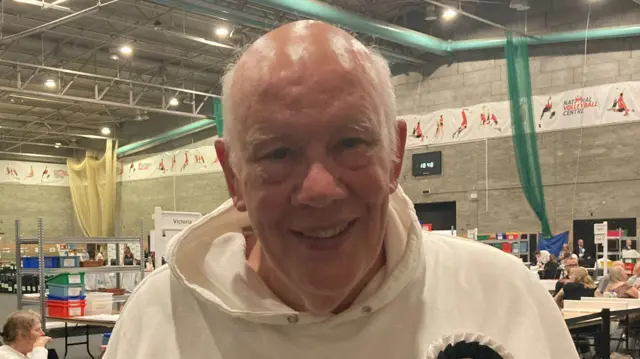 Jim Hakewill wearing a white hoodie, smiling at the election count inside a sports hall.