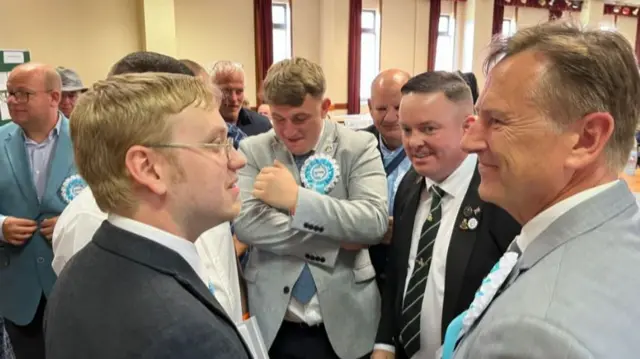 A gathering of men in suits with pale blue rosettes