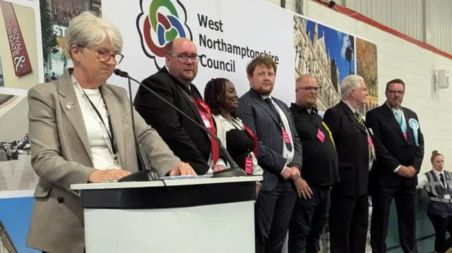 Six candidates wearing smart clothes stand on stage as the council chief executive reads the results from behind a lectern.