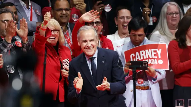 Carney smiling and about to clap, a large crowd of happy people stand around behind him