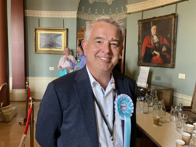 A man with grey hair, wearing a pale blue or white shirt under a blue blazer, smiles at the camera. He is wearing a blue rosette labelled "REFORM UK".