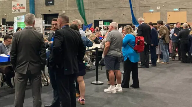 Groups of people watching the votes being counted inside a large sports hall.
