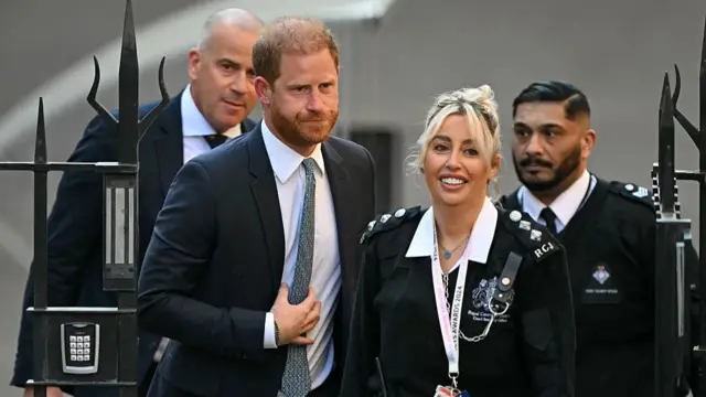 Prince Harry walking through a metal gate surrounded by one female and one male police officer. Another man in a suit stands behind.