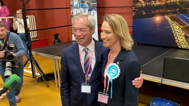 Farage smiles and has his arm around Reform candidate Sarah Pochin who is also smiling as photographers crowd round