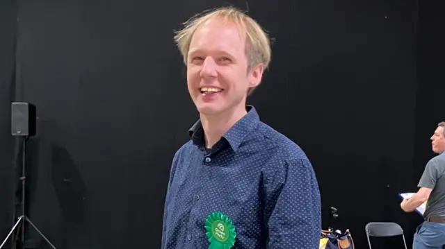 A man in a navy-long sleeve shirt, he is wearing a Green Party rosette and smiling at the camera. There is a black wall behind him.