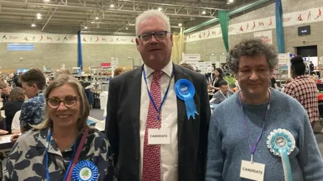Wendy Brackenbury, David Brackenbury and Joseph Garner standing next to each other. They are all dressed smartly while wearing rosettes that are either royal blue or pale blue. They look pleased.