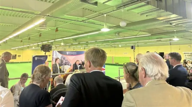 A crowd of people are standing in a bowling centre, watching a podium that is place in front of a Shropshire Council banner