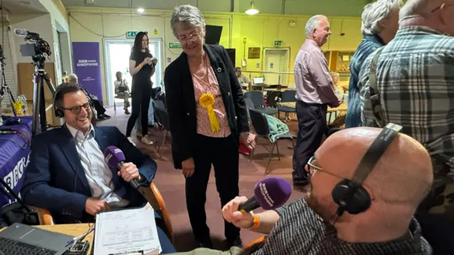 Heather Kidd has grey hair and is talking to two BBC reporters. She has a yellow lib dem rosette on her chest.