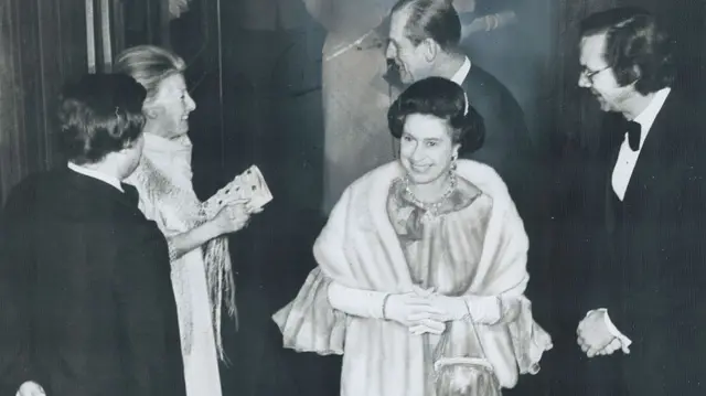 the Queen smiles as she exits a room, her husband is behind her. There's people on either side