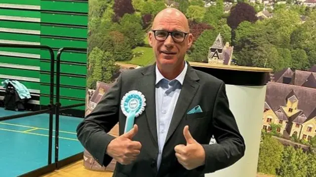 A picture of Anthony Owen, a Reform UK councillor in Hertfordshire. He is wearing a dark suit and a Reform UK rosette, has brown shoes and is giving the camera the thumbs up.