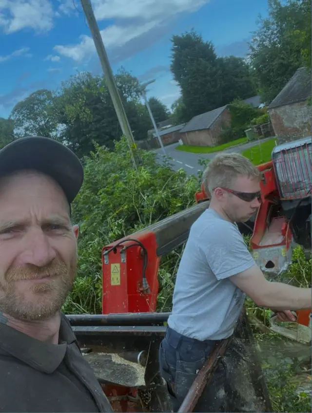 A selfie taken by Daniel Graham shows Daniel Graham in the foreground and Adam Carruthers working behind him. Daniel Graham is looking at the camera and wearing a black cap and black shirt. Adam Carruthers is side on and wearing sunglasses and a grey top
