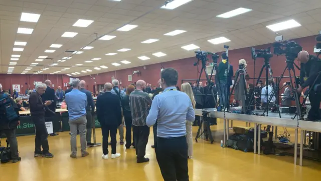 People stand around, and cameras are positioned on a raised platform, as counting continues in the background