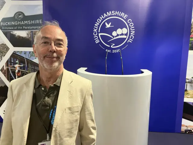 Martin Tett a man with a cream jacket and a khaki shirt. He is looking at the camera and smiling.