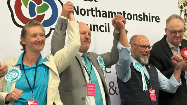 The three successful candidates wear pale blue and lift their arms in the air in celebration