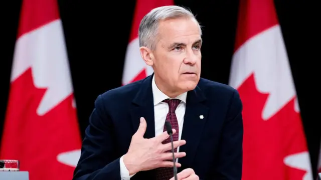Carney holds a hand to his chest as he speaks into a microphone while sitting down
