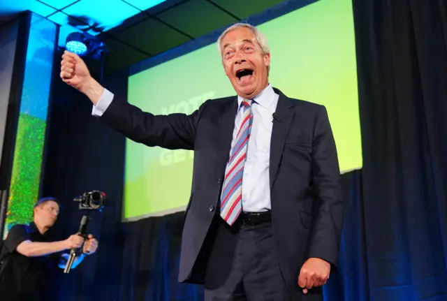 Nigel Farage is on a stage with a wide smile and his arm outstretched. He is wearing a black suit and striped tie