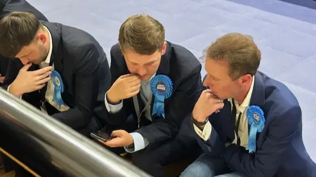 Three men wearing Tory rosettes look nervous as they sit and wait for the result