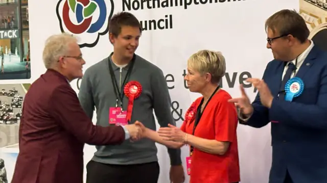 Ian McCord shakes hands with a fellow candidate on stage, who is wearing red. He is clapped by another man having won the seat.