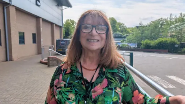 A woman with long ginger hair and a green and black floral top standing beside a metal railing outside a large building