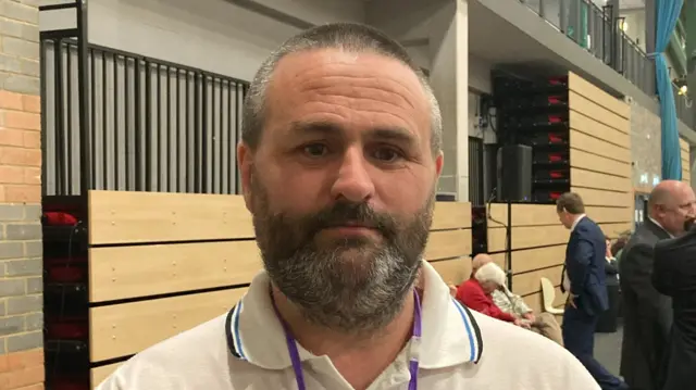 Trevor Conway looking serious while wearing a white polo shirt in the sports hall.