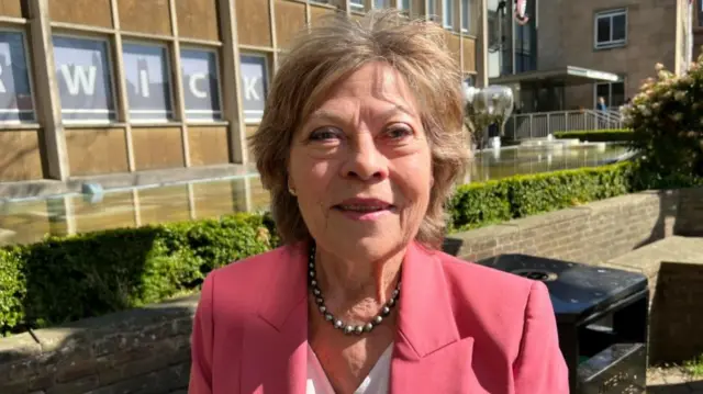 A woman in a pink blazer outside of a building. She is smiling and has short brown hair. The sun is shining behind her.