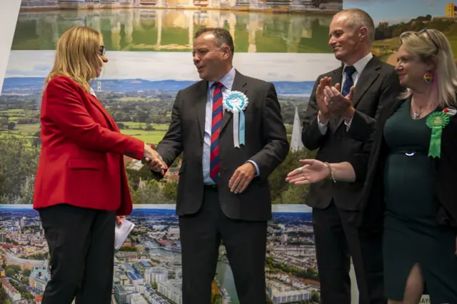 Four political candidates shaking eachother's hands after an election result. A woman is wearing a red blazer while shaking a man's hand who is wearing a suit and a tie.