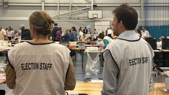 Two people in a sports village with white bibs on that say "election staff" on the back. There are lots of wooden tables in the background with people sitting and standing nearby