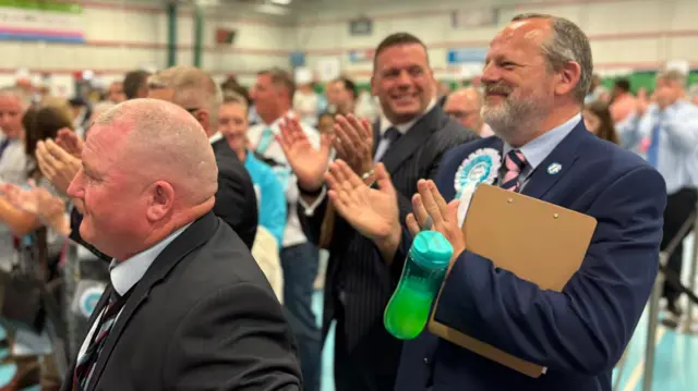 Three Reform UK campaigners wearing suits and pale rosettes clapping.