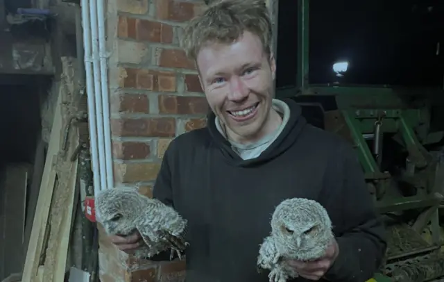 Adam Carruthers holding two owls. He is holding one in each hand and is wearing a black hoodie and smiling at the camera