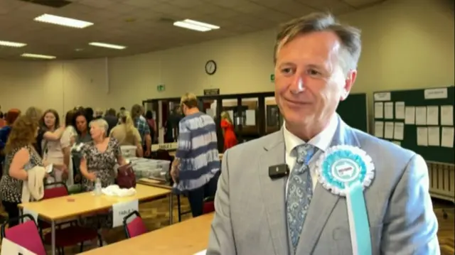 A man in a grey suit in a crowded room with a pale blue rosette