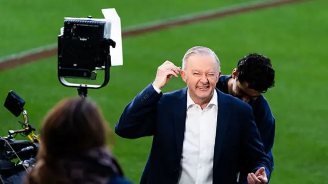 Prime Minister of Australia Anthony Albanese prepares to do live TV interviews at the MCG.  He can be seen putting in an earpiece in front of a TV camera.