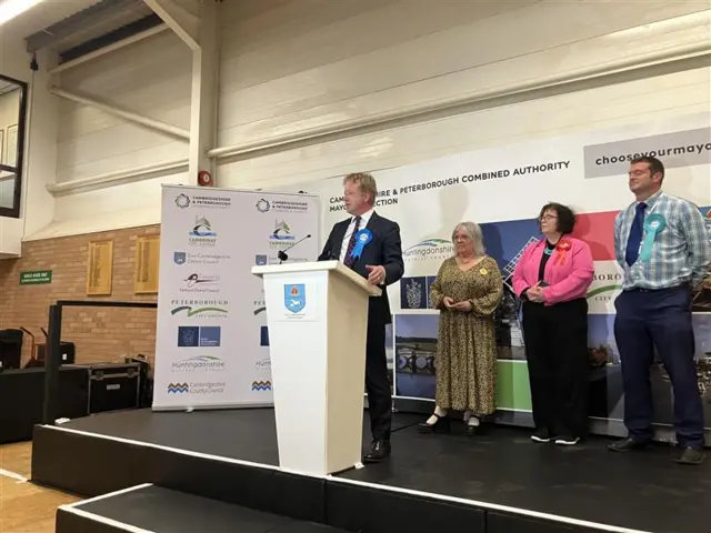 Paul Bristow stands at a lectern giving a speech after winning the election. Three candidates stand behind him. He is wearing a suit and blue rosette.