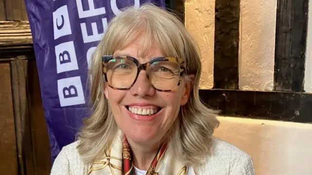 A woman with shoulder-length blonde hair and glasses with patterned frames is wearing a white jacket and a patterned shawl.
