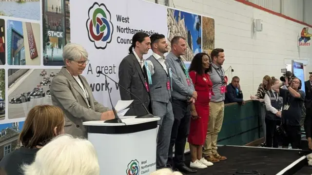 Five candidates wearing smart clothes stand on a stage at the West Northants count. The council chief executive is reading the results from behind a lectern.