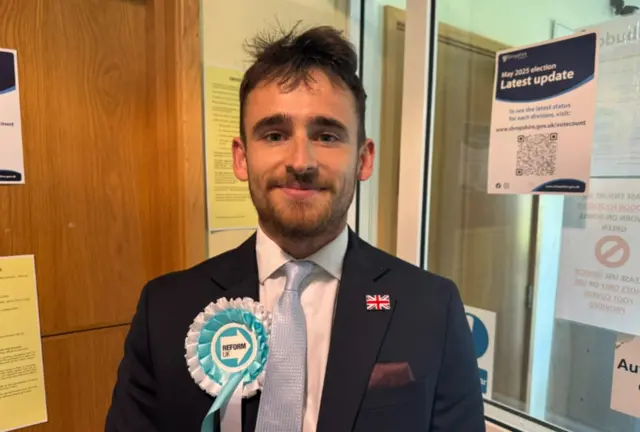 A man with dark ruffled hair and beard, and a Reform UK rosette on his black suit