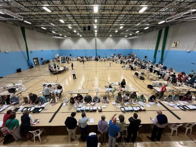 A sports hall with people in counting votes whilst others watch on, monitoring the counting process