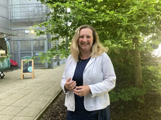 Lucy Nethsingha is smiling at the camera while standing outside. She is wearing a white blouse and blue dress.