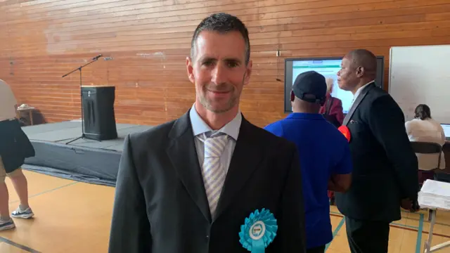 A man in a suit with a pale blue Reform rosette. He is standing in a sports hall and smiling.