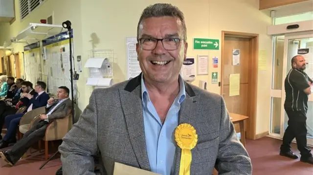 Andy Hall has short dark and grey hair and wears glasses. He is wearing a grey suit with a yellow liberal democrats rosette on his chest
