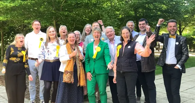 Pippa Heylings in a green suit cheers alongside a group of councillors as they celebrate their victories. Many of them are wearing yellow Liberal Democrat rosettes.