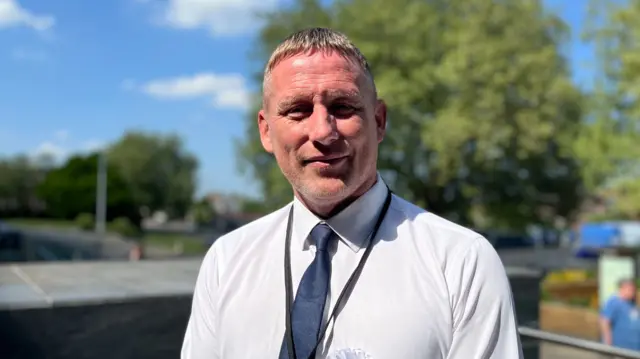 Chris Vaughan looks at the camera in a shirt and tie with reform rossette.