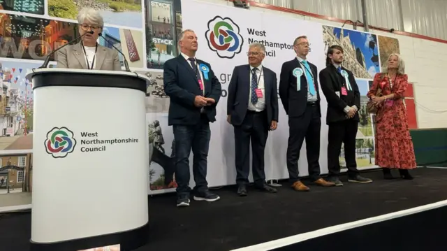 Five smartly dressed people on stage as the results are read out by a woman standing behind a lectern.