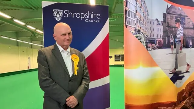 John Tandy is bald and is wearing a dark suit with a yellow Lib Dem rosette badge