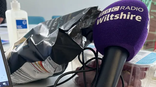 A mic balanced on a pack of grapes and a bag of crisps next to a laptop