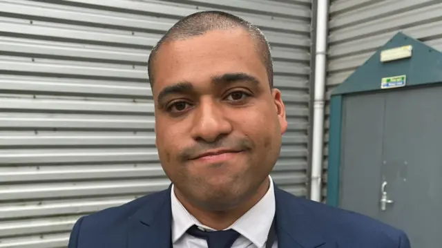James Hill looking glum outside a grey building. He is wearing a white shirt, navy tie and navy suit jacket.