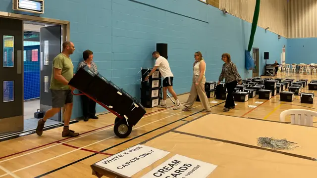 Inside a big sports hall are black ballot boxes, including one being wheeled in. Several other people are helping.